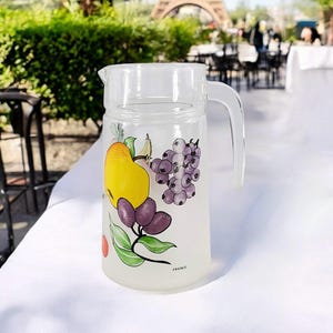 May include: A clear glass jug with a frosted finish and a handle. The jug features a colourful design of fruit and berries, including a yellow pear, purple grapes, and blue berries. The text "France" is printed on the base of the jug.