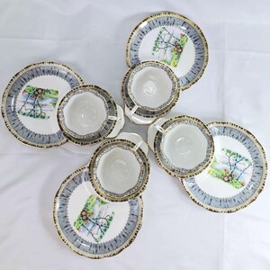 Puede incluir: Juego de t&eacute; de seis piezas con tazas y platillos blancos, borde azul y dorado. Cada platillo presenta un dise&ntilde;o de paisaje. El juego est&aacute; dispuesto sobre un fondo blanco.