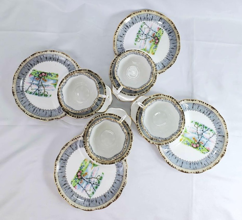 Puede incluir: Juego de t&eacute; de seis piezas con tazas y platillos blancos, borde azul y dorado. Cada platillo presenta un dise&ntilde;o de paisaje. El juego est&aacute; dispuesto sobre un fondo blanco.