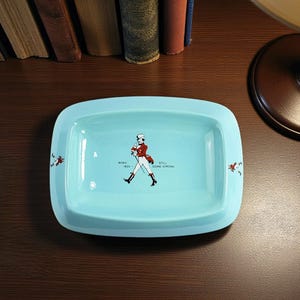 May include: A rectangular, pale blue ceramic tray with rounded corners. It features a central illustration of a man in a red coat and black boots, with the text "BORN 1820 - STILL GOING STRONG". Small figures are on the corners.