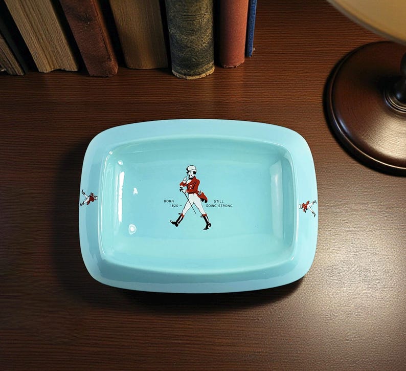 May include: A rectangular, light blue ceramic tray with rounded corners. The tray features a central illustration of a man in a red coat and black boots, with the text "BORN 1820 - STILL GOING STRONG". Small figures are on the corners.
