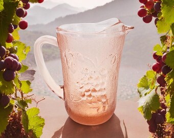 May include: A pink glass pitcher with a grape design. The pitcher has a textured surface and a handle. The pitcher is sitting on a table with a blurred background of a vineyard.