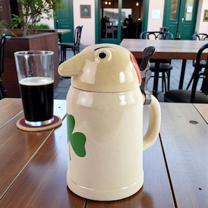 Peut inclure: Une chope à bière en céramique avec un couvercle fantaisiste en forme de nez et un trèfle vert. La chope beige a une poignée et un fermoir en métal. Un verre de bière noire est posé sur un dessous de verre à côté de la chope.