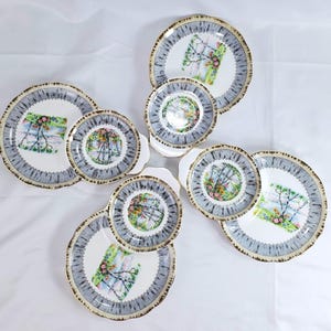 Puede incluir: Un juego de platos y platillos de porcelana vintage. Los platos tienen un centro blanco con un dise&ntilde;o floral y paisaj&iacute;stico colorido. Los bordes son grises con un ribete dorado. El juego incluye platos grandes y platillos m&aacute;s peque&ntilde;os.