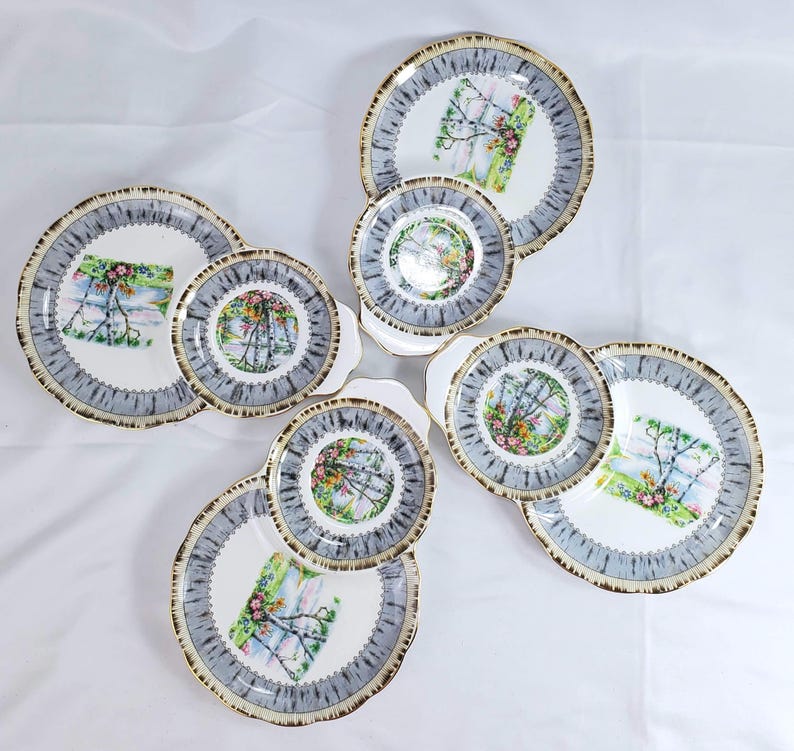 Puede incluir: Un juego de platos y platillos de porcelana vintage. Los platos tienen un centro blanco con un dise&ntilde;o floral y paisaj&iacute;stico colorido. Los bordes son grises con un ribete dorado. El juego incluye platos grandes y platillos m&aacute;s peque&ntilde;os.