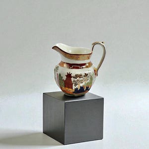 May include: A small, antique cream jug with a detailed landscape design. The jug has a white interior, a brown and gold rim, and a curved handle. The exterior features a colourful scene with trees, buildings, and figures, set against a white background.
