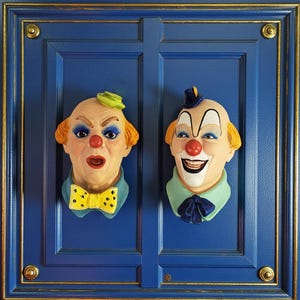 Peut inclure: Deux décorations murales en forme de tête de clown. L'un a un chapeau vert et un nœud papillon jaune, l'autre un chapeau bleu et un nœud papillon. Les deux ont des nez rouges, des visages peints et des cheveux orange. Les clowns sont montés sur une porte bleue avec des bordures dorées.