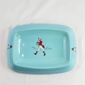 May include: A rectangular, light blue ceramic tray with rounded corners. The tray features a central illustration of a man in a red coat and black boots, with the text "BORN 1820 - STILL GOING STRONG". Two smaller figures are on the sides.