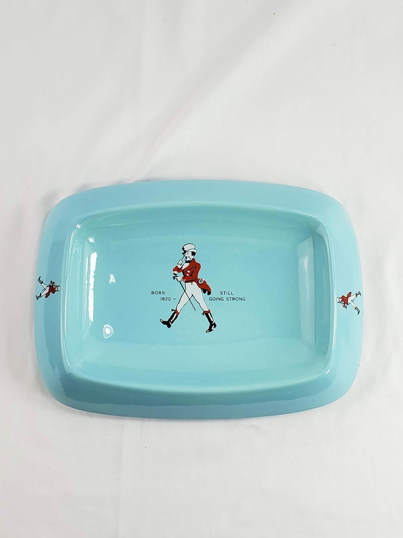 May include: A rectangular, light blue ceramic tray with rounded corners. The tray features a central illustration of a man in a red coat and black boots, with the text "BORN 1820 - STILL GOING STRONG". Two smaller figures are on the sides.