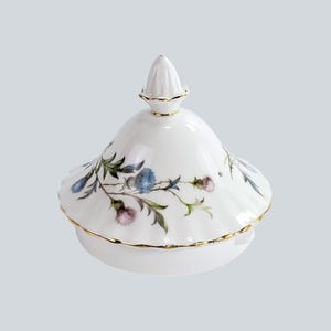 Può includere: Coperchio in porcellana bianca con finiture dorate e un motivo floreale di cardi blu e viola. Il coperchio ha un terminale conico e un bordo smerlato. Coperchio decorativo per teiera o piatto da portata.