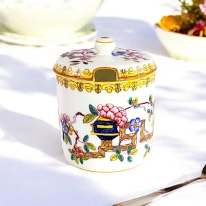Peut inclure: Un sucrier en porcelaine blanche avec un couvercle, orné d'un motif floral dans des tons de rose, bleu, jaune et vert. Le bol est bordé d'or et comporte une petite ouverture dans le couvercle pour verser.