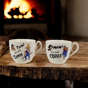 Peut inclure: Deux tasses à café en céramique blanche avec des illustrations de dessins animés et le texte "Paw! Git your coffee" et "Maw! Git your coffee".