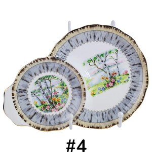 Puede incluir: Dos platos decorativos de porcelana con una escena de paisaje. Los platos tienen un borde gris y dorado con una escena de paisaje de &aacute;rboles, flores y una masa de agua. Los platos se exhiben en colgadores de platos blancos.