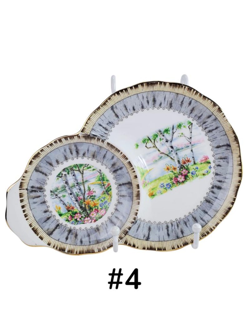 Puede incluir: Dos platos decorativos de porcelana con una escena de paisaje. Los platos tienen un borde gris y dorado con una escena de paisaje de &aacute;rboles, flores y una masa de agua. Los platos se exhiben en colgadores de platos blancos.