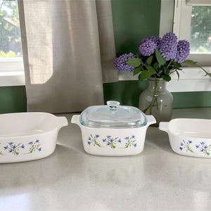 May include: A collection of three white ceramic oven dishes with blue floral patterns. One dish has a glass lid. The dishes are on a white surface, with a vase of purple flowers in the background.
