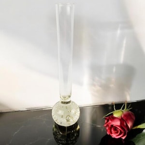 May include: A clear glass vase with a round, textured base. The vase is tall and slender, with a simple design. The base of the vase is made of clear glass with small air bubbles throughout.