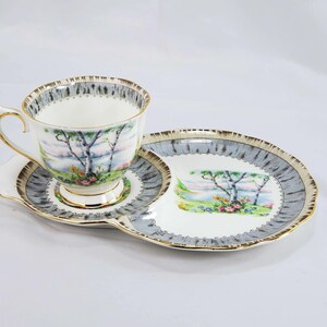 Puede incluir: Juego de t&eacute; vintage con taza, platillo y plato de servir. El juego presenta una escena de paisaje con &aacute;rboles y flores, enmarcada por un borde gris y dorado. La taza y el plato tienen ribete dorado.