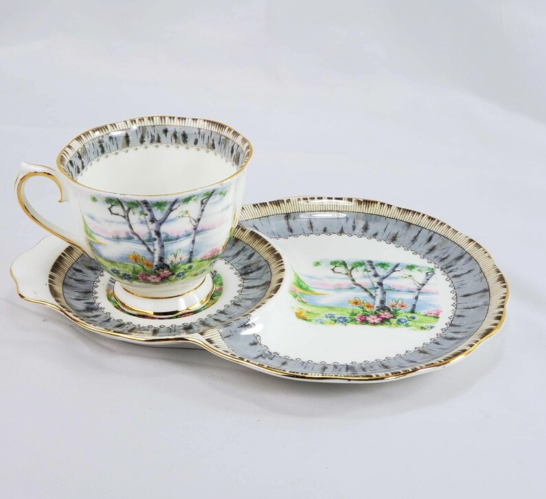 Puede incluir: Juego de t&eacute; vintage con taza, platillo y plato de servir. El juego presenta una escena de paisaje con &aacute;rboles y flores, enmarcada por un borde gris y dorado. La taza y el plato tienen ribete dorado.