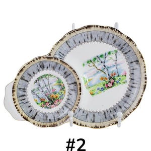 Puede incluir: Plato de servir vintage de dos piezas con un dise&ntilde;o de paisaje. Los platos tienen un borde gris y marr&oacute;n con un interior blanco. El centro de los platos presenta un paisaje colorido con &aacute;rboles, flores y agua.
