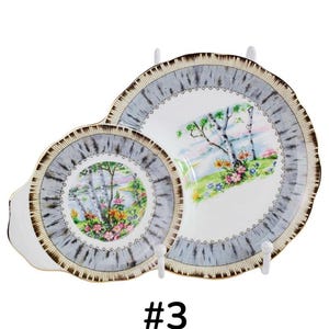 Puede incluir: Juego de servir de porcelana vintage de dos piezas. Los platos presentan una escena de paisaje con &aacute;rboles y flores. Los bordes tienen un patr&oacute;n gris y marr&oacute;n. El juego se exhibe en soportes blancos.