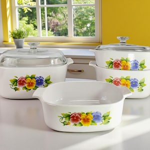 May include: A collection of white ceramic cookware featuring a pansy floral pattern. The set comprises several casserole dishes and a serving dish, all with handles. The pansies are red, yellow, and blue.