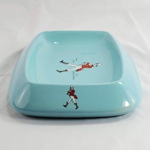 May include: A light blue ceramic ashtray with rounded edges. The ashtray features a graphic of a man in a red coat and the text "STILL GOING STRONG". The design includes the years "1820 - 1923".