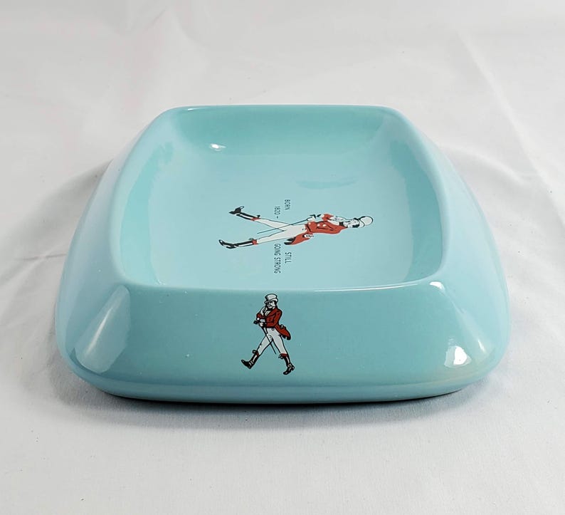 May include: A light blue ceramic ashtray with rounded edges. The ashtray features a graphic of a man in a red coat and the text "STILL GOING STRONG". The design includes the years "1820 - 1923".