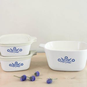 May include: A set of white ceramic baking dishes featuring blue floral patterns. The collection comprises two rectangular dishes with lids and a larger square dish. They are displayed on a light wooden surface, with a few blue flowers.