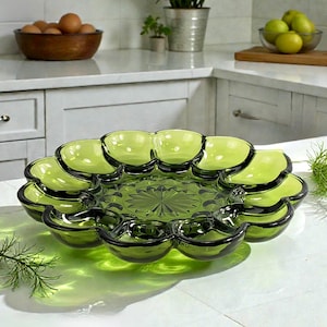 May include: A green glass serving dish with multiple compartments, arranged in a flower-like shape. The dish has a central, patterned base and dark green rims. The dish is on a white surface.
