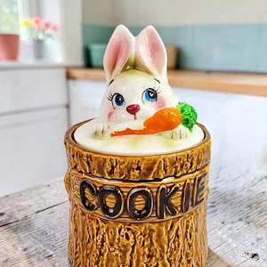 以下が含まれることがあります： 茶色の質感のある切り株の形をした陶器製のクッキー瓶。側面には濃い茶色の文字で「COOKIE」と刻まれています。蓋はピンクの耳、青い目、オレンジ色のニンジンを持った白いウサギの形をしています。