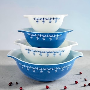 Peut inclure: Une pile de quatre saladiers Pyrex de différentes tailles. Les bols alternent entre le blanc et le bleu, chacun orné d'un motif de flocons de neige et de points blancs. Les bols ont des poignées et sont disposés sur une surface grise avec des baies rouges.