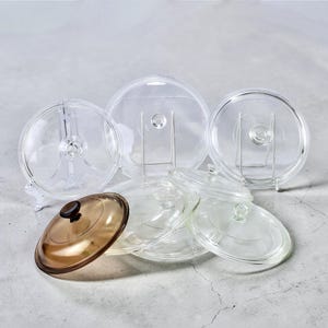 May include: A selection of clear glass pot lids, some with metal stands. One lid is amber-coloured with a black knob. The lids vary in size and shape, suitable for various cookware.
