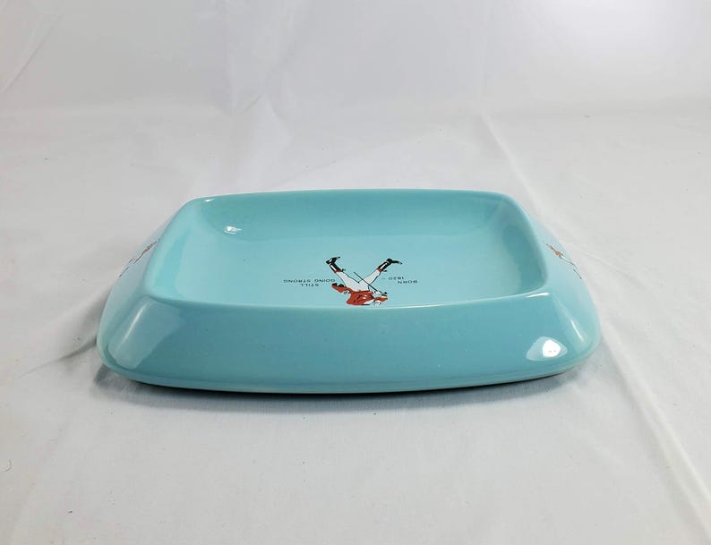May include: A rectangular, light blue ceramic ashtray. The ashtray has rounded corners and a slightly raised edge. The center of the ashtray features a graphic of a skier. Text on the ashtray reads "Colorado Rockies" and "Ski Team".