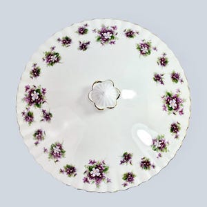 May include: A white, round serving dish lid with a scalloped edge and a floral pattern of purple and white violets. The lid has a white, flower-shaped handle with gold trim. The rim of the lid is also trimmed in gold.