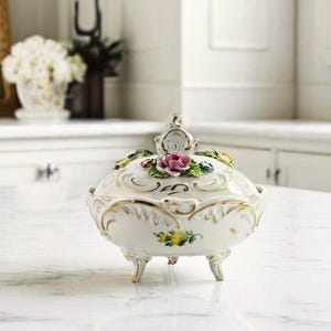 May include: A white porcelain trinket box with a lid, featuring floral accents and gold trim. The box has three small feet and a decorative handle on top. The lid is adorned with a pink rose and other colourful flowers.