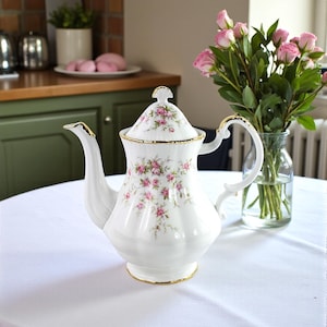 May include: A white porcelain teapot with a floral design of pink roses and green foliage. The teapot features gold edging and a matching lid. A vase of pink roses stands close by. The teapot is placed on a white tablecloth.