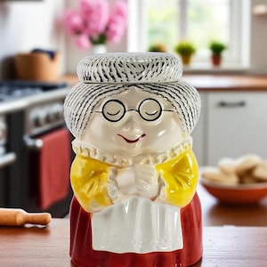 以下が含まれることがあります： 笑顔のおばあちゃんの形をした陶器のクッキーの瓶。瓶は、黒いストライプの白い帽子、丸いメガネ、赤いドレスが特徴です。黄色の袖と白いエプロンが付いています。