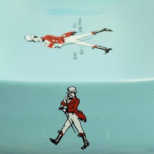 May include: A light blue ceramic ashtray featuring two illustrations of a man in a red coat and white pants. One illustration is walking, and the other is lying down. Text includes "BORN 1820" and "STILL GOING STRONG."