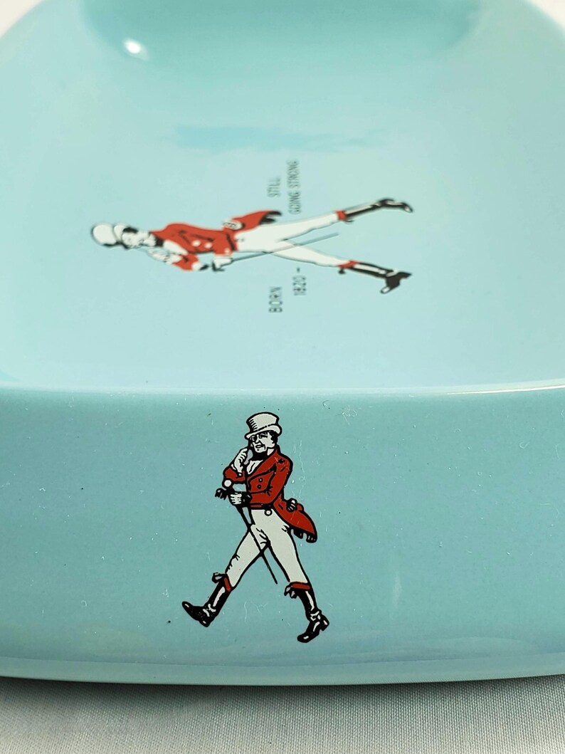 May include: A light blue ceramic ashtray featuring two illustrations of a man in a red coat and white pants. One illustration is walking, and the other is lying down. Text includes "BORN 1820" and "STILL GOING STRONG."