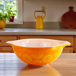 Peut inclure: Un saladier orange avec un intérieur blanc et un motif de marguerites. Le saladier est en verre et possède deux poignées. Le saladier est posé sur une table en bois. D'autres articles de cuisine sont en arrière-plan.