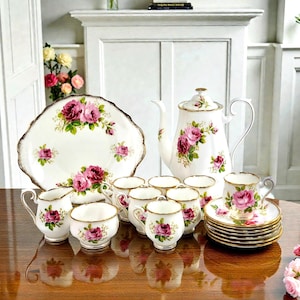 Könnte beinhalten: Ein Vintage-Teeservice mit weißer Porzellan-Teekanne, Tassen, Untertassen, Sahnekännchen, Zuckerdose und Servierplatte. Jedes Stück ist mit rosa Rosenmustern und goldenen Verzierungen versehen, was eine elegante Ausstrahlung erzeugt.