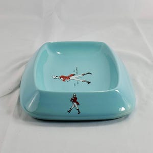 May include: A light blue ceramic ashtray with a square shape and rounded edges. The ashtray features a graphic of a person in a red coat and black pants. The text "STILL GOING STRONG" is also visible.