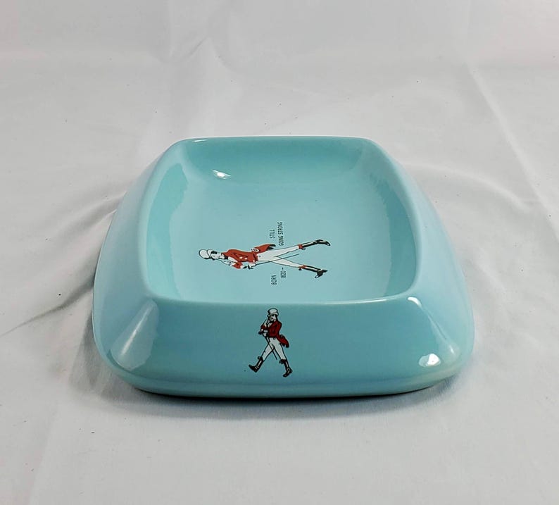 May include: A light blue ceramic ashtray with a square shape and rounded edges. The ashtray features a graphic of a person in a red coat and black pants. The text "STILL GOING STRONG" is also visible.