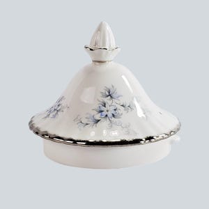 May include: White porcelain lid with a silver rim and a conical finial. The lid is decorated with blue floral patterns. A decorative piece for a tea set or similar items.