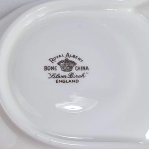 Puede incluir: Una fuente blanca de porcelana Royal Albert Bone China con forma curva. La fuente presenta el patr&oacute;n "Silver Birch" y las palabras "Royal Albert Bone China England" en letras marrones.