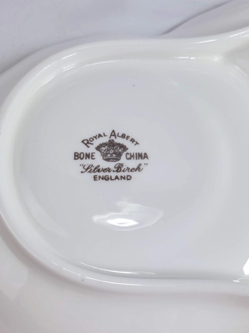 Puede incluir: Una fuente blanca de porcelana Royal Albert Bone China con forma curva. La fuente presenta el patr&oacute;n "Silver Birch" y las palabras "Royal Albert Bone China England" en letras marrones.