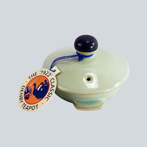 Puede incluir: Tapa de tetera de cerámica verde pálido con un pomo negro y un pequeño agujero. Una etiqueta adjunta con una cuerda azul dice "THE 1922 CLASSIC DENBY TEAPOT" con una ilustración de tetera. La tapa tiene forma redondeada.