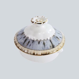 May include: A white ceramic serving dish with a lid. The lid has a light blue and grey pattern with gold trim. The lid features a small, round handle with gold accents. The dish is likely for serving food.