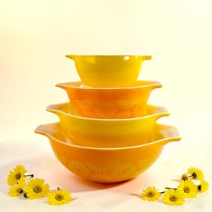 Peut inclure: Ensemble de quatre bols à mélanger Pyrex vintage, dans des tons de jaune et d'orange. Les bols présentent un motif floral et des poignées. Des marguerites jaunes sont disposées autour de la base des bols.