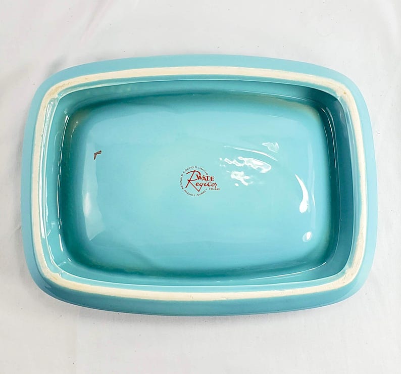 May include: A rectangular, turquoise ceramic dish with a white rim. The interior is a solid turquoise color. The dish has rounded corners and a slightly raised edge. The bottom of the dish has a red and white logo that reads "Wade Regicor".